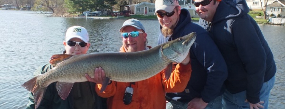 Four people standing by a lake, one holding a large fish horizontally and the others smiling. Trees and houses are in the background.